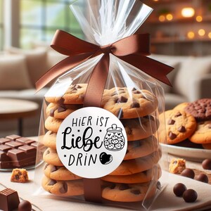 May include: A clear plastic bag filled with chocolate chip cookies tied with a brown ribbon. A white sticker on the bag reads "Hier ist Liebe drin" with a black and white illustration of a jar filled with cookies.
