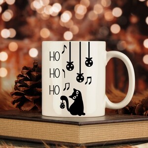 May include: White ceramic mug with a black silhouette of a cat holding a Christmas ornament. The mug has the words "Ho Ho Ho" written on it with musical notes and hanging ornaments.