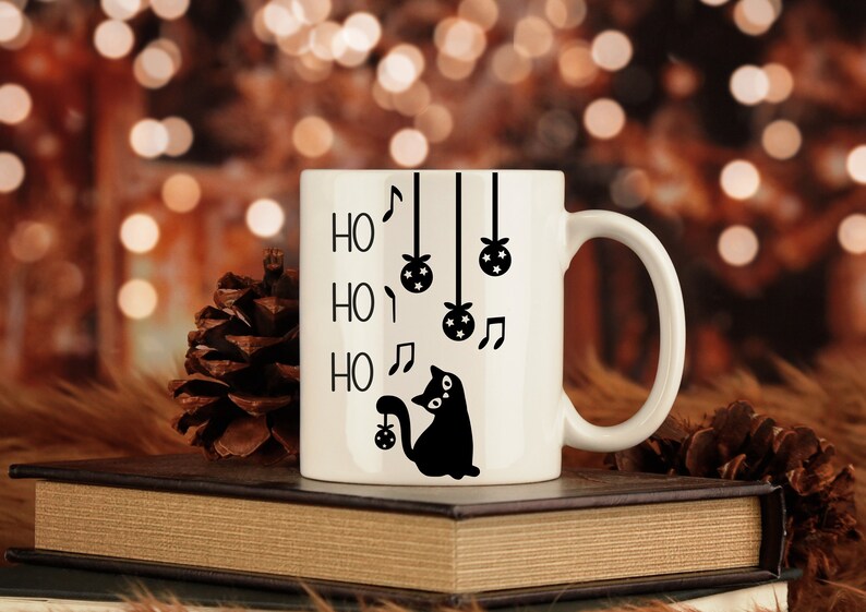 May include: White ceramic mug with a black silhouette of a cat holding a Christmas ornament. The mug has the words "Ho Ho Ho" written on it with musical notes and hanging ornaments.