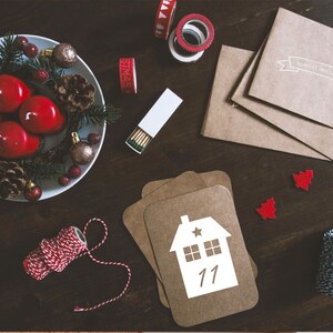 K&ouml;nnte beinhalten: Ein Adventskalender aus braunem Papier mit einem wei&szlig;en Hausdesign und der Nummer 11. Der Kalender liegt auf einem Holztisch mit einem Kranz aus roten Kerzen, Tannenzweigen und Tannenzapfen. Au&szlig;erdem gibt es rotes und wei&szlig;es Garn, Streichh&ouml;lzer und einen wei&szlig;en Umschlag mit dem Text "Special Delivery for you".