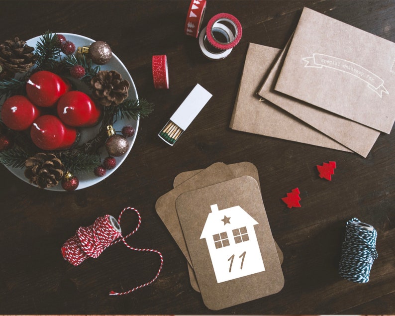 K&ouml;nnte beinhalten: Ein Adventskalender aus braunem Papier mit einem wei&szlig;en Hausdesign und der Nummer 11. Der Kalender liegt auf einem Holztisch mit einem Kranz aus roten Kerzen, Tannenzweigen und Tannenzapfen. Au&szlig;erdem gibt es rotes und wei&szlig;es Garn, Streichh&ouml;lzer und einen wei&szlig;en Umschlag mit dem Text "Special Delivery for you".