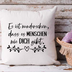 May include: White throw pillow with black text that reads "Es ist wundersch&ouml;n, dass es Menschen wie DICH gibt."  The text is surrounded by two small floral designs with a heart in the center.