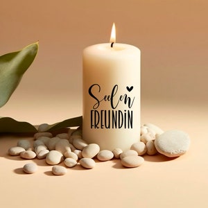 Puede incluir: Una vela blanca con el texto "Seelen Freundin" y un peque&ntilde;o coraz&oacute;n en el lateral. La vela est&aacute; rodeada de piedras blancas y una hoja verde.
