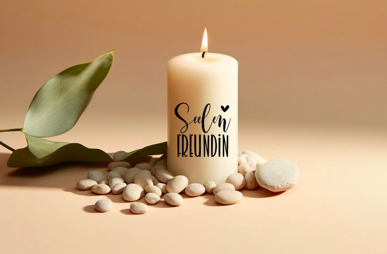 Puede incluir: Una vela blanca con el texto "Seelen Freundin" y un peque&ntilde;o coraz&oacute;n en el lateral. La vela est&aacute; rodeada de piedras blancas y una hoja verde.