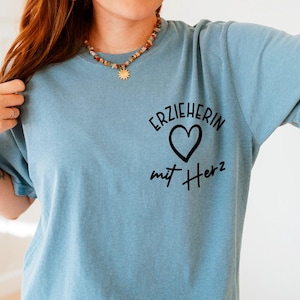 May include: A light blue t-shirt with the German text "ERZIEHERIN mit Herz" printed in black. The design includes a heart symbol. The shirt is made of a soft material.