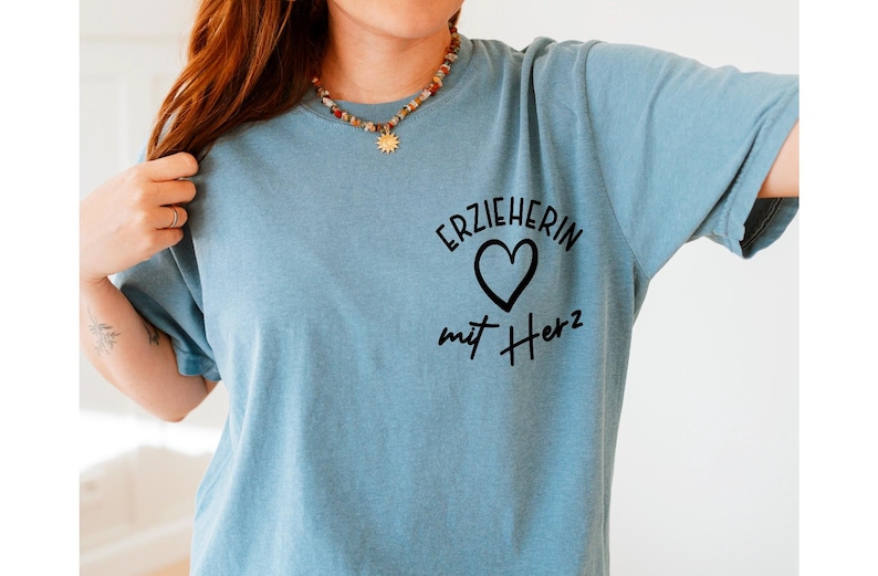 May include: A light blue t-shirt with the German text "ERZIEHERIN mit Herz" printed in black. The design includes a heart symbol. The shirt is made of a soft material.