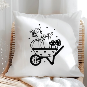 Op de afbeelding: Een wit kussen met een zwart-witte illustratie van een kruiwagen gevuld met pompoenen en paddenstoelen. Een klein vogeltje zit bovenop de pompoenen.