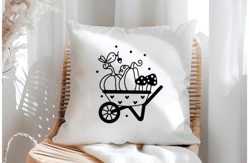 Op de afbeelding: Een wit kussen met een zwart-witte illustratie van een kruiwagen gevuld met pompoenen en paddenstoelen. Een klein vogeltje zit bovenop de pompoenen.