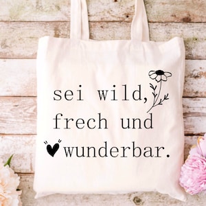 May include: A white tote bag with black text that reads "sei wild, frech und wunderbar."  A small black heart is above the word "wunderbar." A black and white flower is to the right of the text.