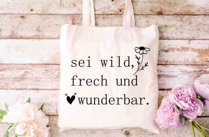 May include: A white tote bag with black text that reads "sei wild, frech und wunderbar."  A small black heart is above the word "wunderbar." A black and white flower is to the right of the text.