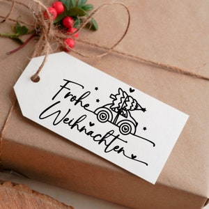 May include: A white gift tag with black text that says "Frohe Weihnachten" and a black and white illustration of a car carrying a Christmas tree.