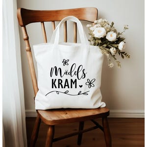 May include: White tote bag with black text that reads "M&auml;ddels Kram" with a small heart and flower designs.