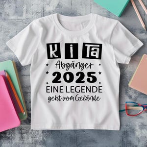 May include: White t-shirt with black text that reads "KiTa Abg&auml;nger 2025 EINE LEGENDE geht vom Gel&auml;nde." The shirt has a crew neck and short sleeves. The text is surrounded by stars.