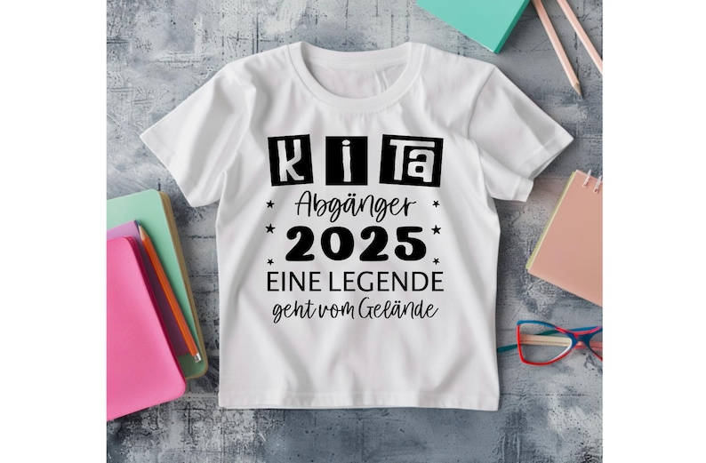 May include: White t-shirt with black text that reads "KiTa Abg&auml;nger 2025 EINE LEGENDE geht vom Gel&auml;nde." The shirt has a crew neck and short sleeves. The text is surrounded by stars.