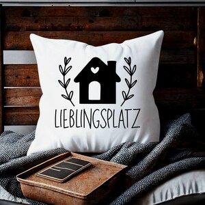 May include: White throw pillow with black text and a black silhouette of a house with a heart on the front. The text reads "Lieblingsplatz".