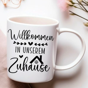 May include: White ceramic mug with a black design that says "Willkommen in unserem Zuhause" with a small house icon and hearts.