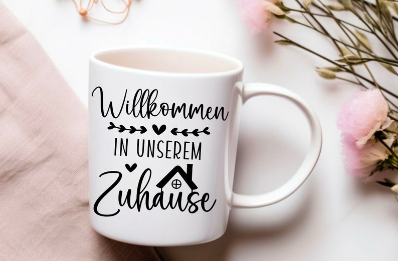 May include: White ceramic mug with a black design that says "Willkommen in unserem Zuhause" with a small house icon and hearts.
