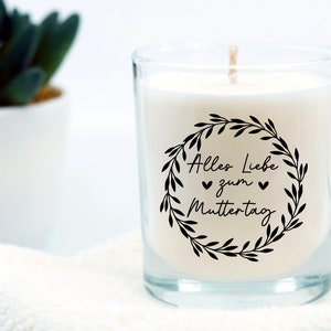 May include: A white candle in a clear glass jar with a black and white label that says "Alles Liebe zum Muttertag" with a wreath of leaves around the text.