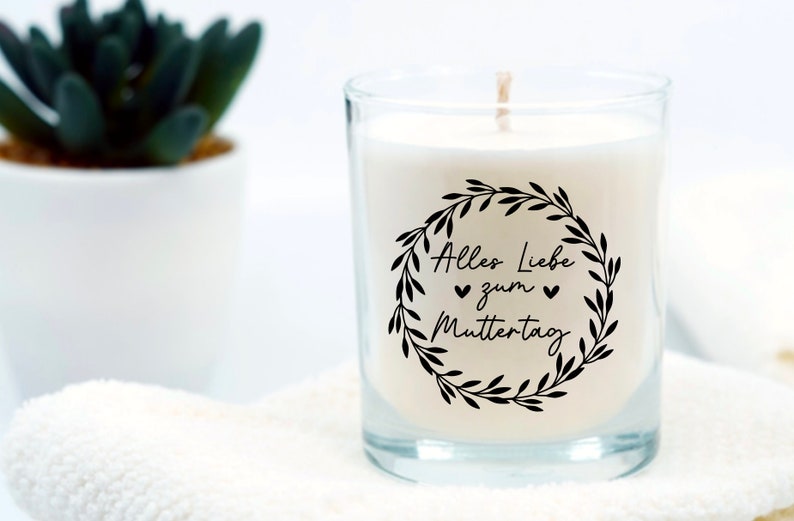 May include: A white candle in a clear glass jar with a black and white label that says "Alles Liebe zum Muttertag" with a wreath of leaves around the text.