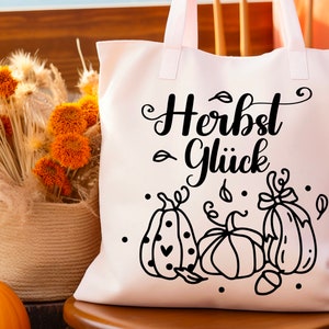 May include: White tote bag with black text that reads "Herbst Gl&uuml;ck" and a black and white illustration of three pumpkins with leaves and dots.