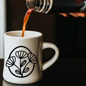 May include: A white ceramic mug with a black outline of a flower design. Coffee is being poured into the mug from a black thermos.