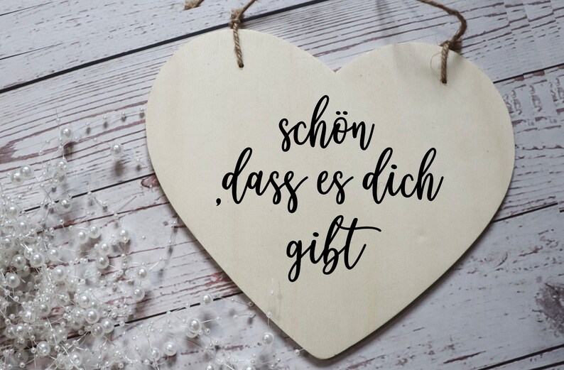 Puede incluir: Un letrero de madera blanco en forma de coraz&oacute;n con el texto "sch&ouml;n dass es dich gibt" en letras cursivas negras.