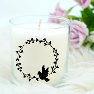 May include: A white candle in a glass jar with a black silhouette of a bird in a wreath design. The candle is on a white fluffy surface.