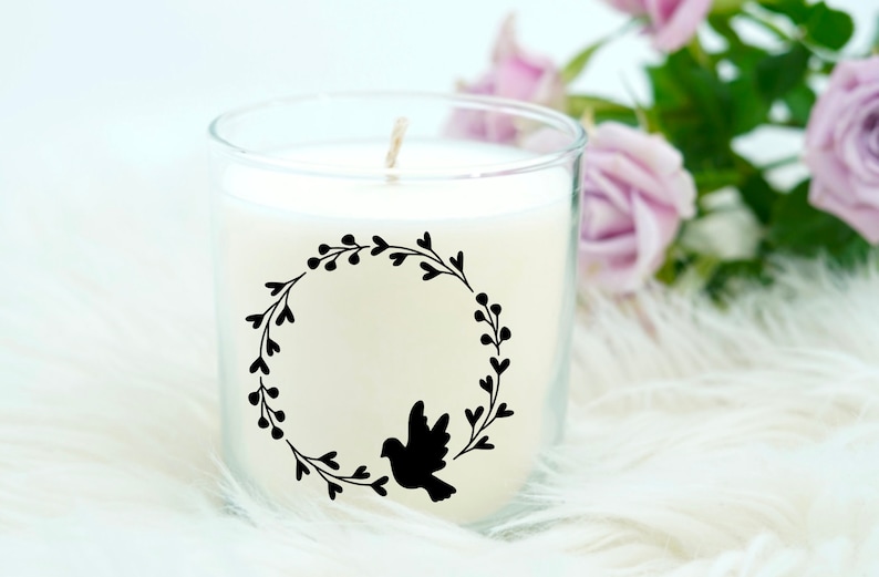 May include: A white candle in a glass jar with a black silhouette of a bird in a wreath design. The candle is on a white fluffy surface.