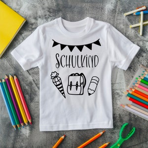 May include: White t-shirt with black text that says "Schulkind" and a black and white illustration of a pencil, backpack, and a cone with a bow on top.