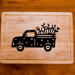 Op de afbeelding: Een zwart-wit houten snijplank met een silhouet van een vrachtwagen die pompoenen vervoert. De vrachtwagen heeft witte stippen en harten op de wielen.