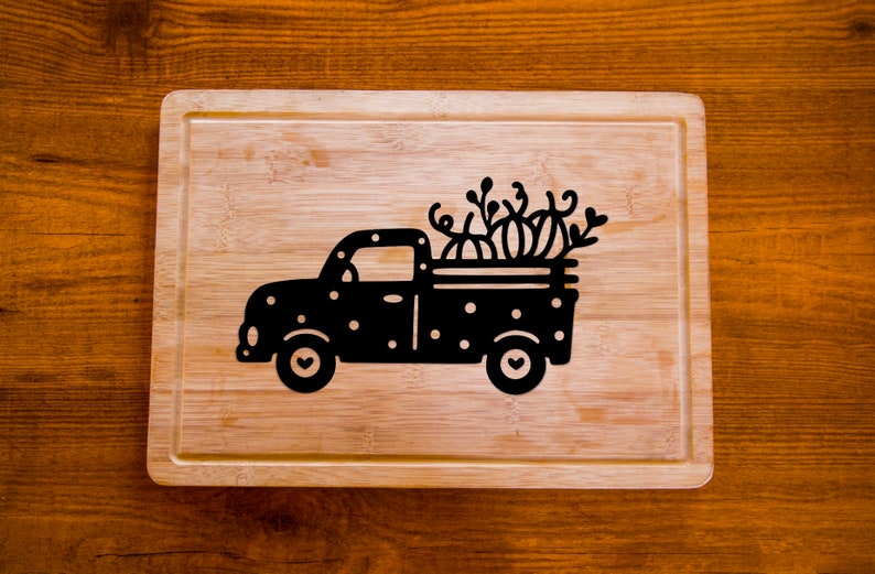 Op de afbeelding: Een zwart-wit houten snijplank met een silhouet van een vrachtwagen die pompoenen vervoert. De vrachtwagen heeft witte stippen en harten op de wielen.
