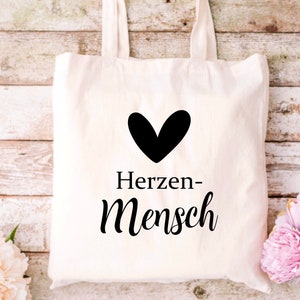 Puede incluir: Una bolsa de tela blanca con texto negro y un coraz&oacute;n negro que dice "Herzen-Mensch".
