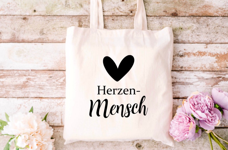 Puede incluir: Una bolsa de tela blanca con texto negro y un coraz&oacute;n negro que dice "Herzen-Mensch".