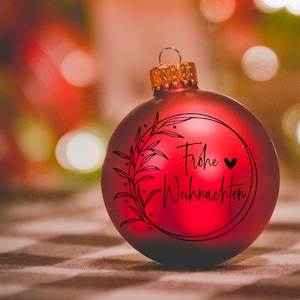 May include: A red Christmas bauble with a black wreath and the text "Frohe Weihnachten" in German. The bauble has a small black heart next to the text.