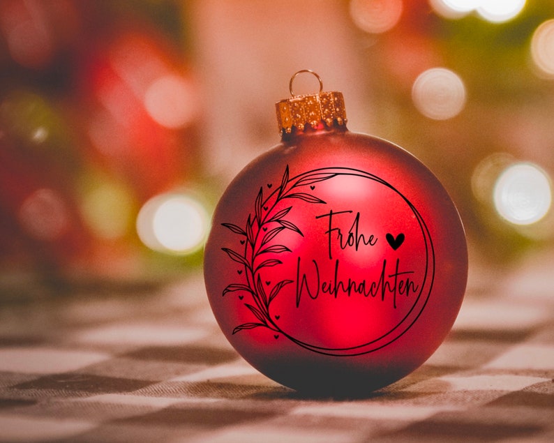 May include: A red Christmas bauble with a black wreath and the text "Frohe Weihnachten" in German. The bauble has a small black heart next to the text.