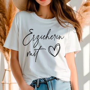 May include: White t-shirt with the German text "Erzieherin mit" and a heart graphic in black. The shirt is a crew neck style and is paired with blue jeans. The image is taken in natural light.
