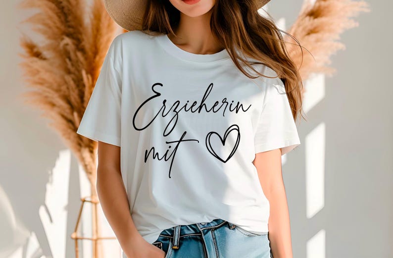 May include: White t-shirt with the German text "Erzieherin mit" and a heart graphic in black. The shirt is a crew neck style and is paired with blue jeans. The image is taken in natural light.