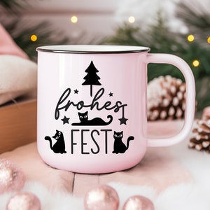 May include: A pink ceramic mug with a black handle and a black design of a Christmas tree, three cats, and the text "frohes FEST".