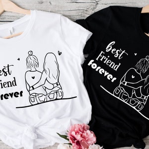 May include: Two white t-shirts with black text and a graphic of two girls hugging. The text on the shirts says "best friend forever".