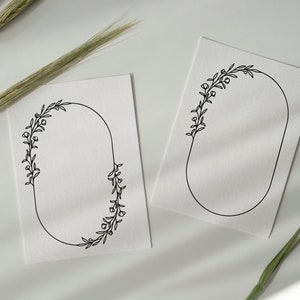 Op de afbeelding: Twee witte kaarten met zwarte, ovale bloemframes. De frames zijn leeg en klaar voor tekst of afbeeldingen. De kaarten liggen op een wit oppervlak met gedroogde tarwehalmen.