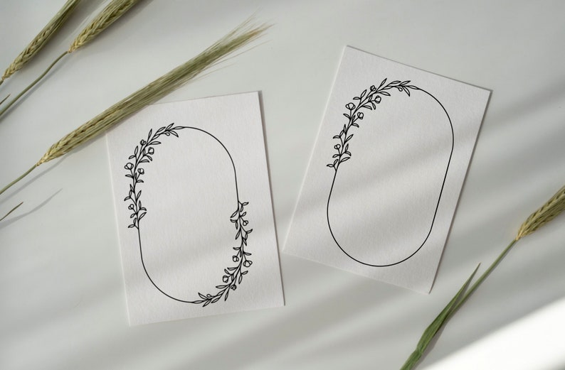 Op de afbeelding: Twee witte kaarten met zwarte, ovale bloemframes. De frames zijn leeg en klaar voor tekst of afbeeldingen. De kaarten liggen op een wit oppervlak met gedroogde tarwehalmen.