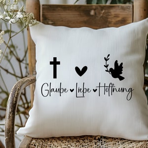 Peut inclure: Un coussin blanc avec du texte et des graphismes noirs. Le texte dit "Glaube Liebe Hoffnung" avec une croix, un c&oelig;ur et une colombe avec une branche d'olivier.
