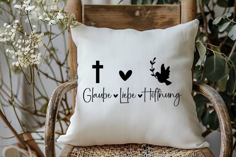 Peut inclure: Un coussin blanc avec du texte et des graphismes noirs. Le texte dit "Glaube Liebe Hoffnung" avec une croix, un c&oelig;ur et une colombe avec une branche d'olivier.