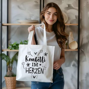 May include: A white tote bag with black text that reads "Konfetti im Herzen" (Confetti in the heart). The bag is being held by a person with long brown hair.