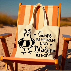 May include: A cream tote bag featuring a cartoon seagull with a life preserver rests on a beach chair. The bag displays the German text "Sonne im Herzen und Bl&ouml;dsinn im Kopf" in black script, with hearts. The chair has orange and white stripes.