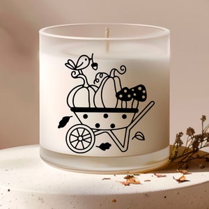 Op de afbeelding: Een witte, gematteerde glazen kaars pot met een zwart-witte illustratie van een kruiwagen gevuld met pompoenen, paddenstoelen en een vogel.