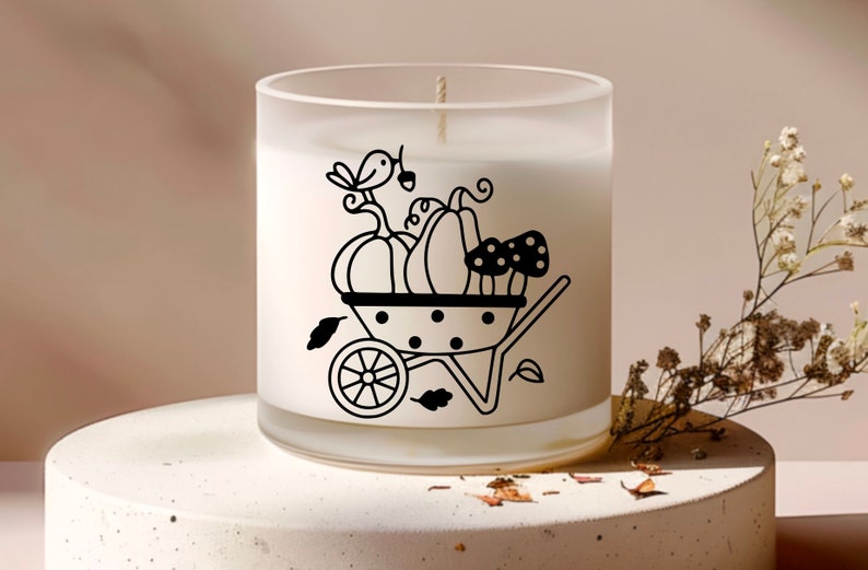 Op de afbeelding: Een witte, gematteerde glazen kaars pot met een zwart-witte illustratie van een kruiwagen gevuld met pompoenen, paddenstoelen en een vogel.