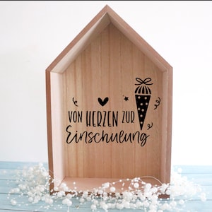 May include: Wooden house-shaped box with a light brown finish. The box features black text and illustrations, including a heart, star, and a school cone. The text reads "VON HERZEN ZUR Einschulung".