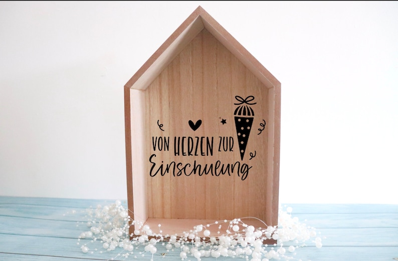 May include: Wooden house-shaped box with a light brown finish. The box features black text and illustrations, including a heart, star, and a school cone. The text reads "VON HERZEN ZUR Einschulung".