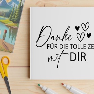 May include: A white card with the German text "Danke f&uuml;r die tolle Zeit mit Dir" in black script, with three heart symbols. The card is on a wooden surface with art supplies and a picture book.